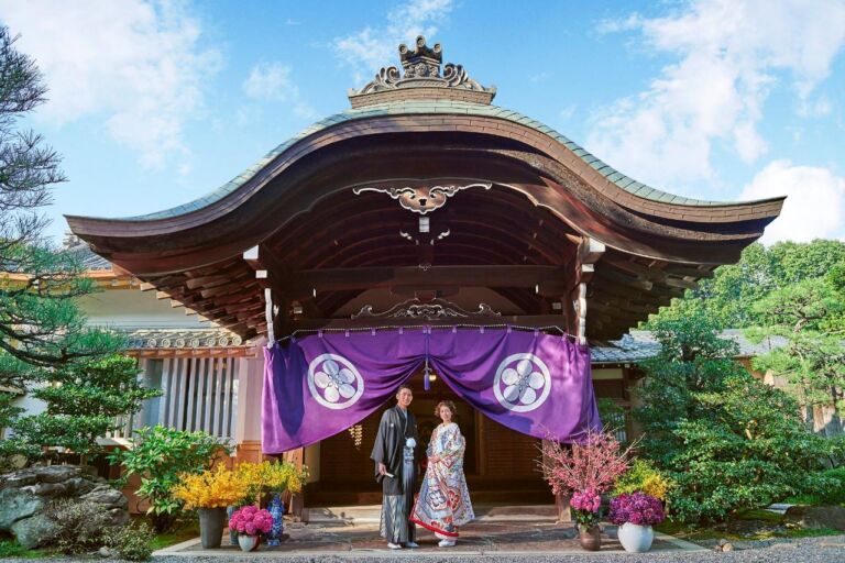 A Japanese couple in traditional attire stands beneath a shrine's ornate roof and purple banner, surrounded by lush greenery and vibrant floral arrangements.