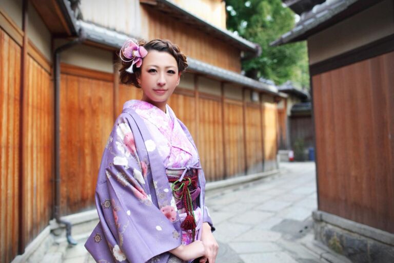 A woman in a purple kimono with pink floral details stands in front of a traditional wooden building in Kyoto, Japan.