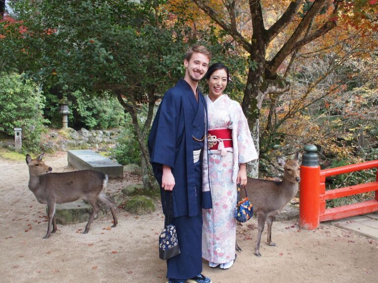 A couple in traditional Japanese clothing pose for a photo with deer in a park-like setting with autumn foliage.