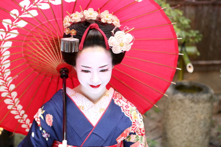 A Japanese woman dressed as a geisha holds a red umbrella and smiles demurely.