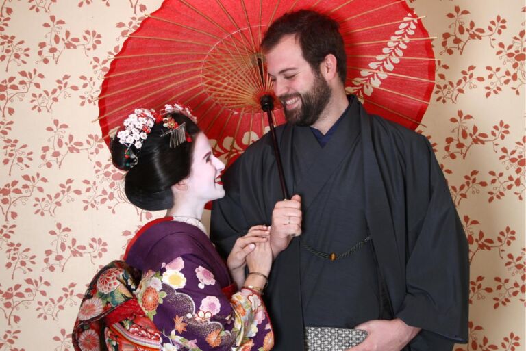 A man and woman dressed in traditional Japanese clothing smile at each other while standing under a red umbrella.