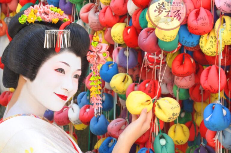 A Geisha in traditional white makeup and ornate hair decorations looks at a collection of colorful fabric charms with handwritten notes in a close-up shot.