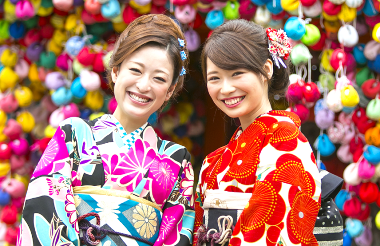 Two young women in vibrant kimonos smile warmly at the camera, standing against a backdrop of colorful hanging charms.