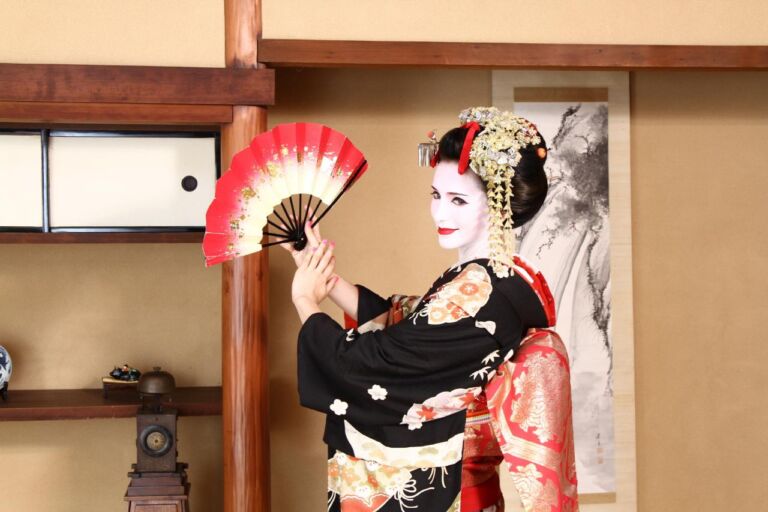 A geisha, with pale white skin and a traditional black kimono, poses with a red and white fan against a backdrop of Japanese architecture.