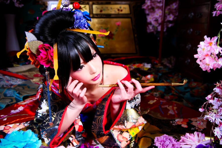 A woman in a traditional Japanese kimono holds a pipe and gazes off-camera with an alluring expression, surrounded by flowers and ornate decorations.