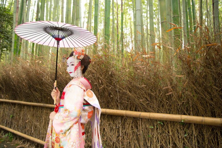 A woman in a traditional Japanese kimono and makeup holds a parasol while standing in a bamboo forest.