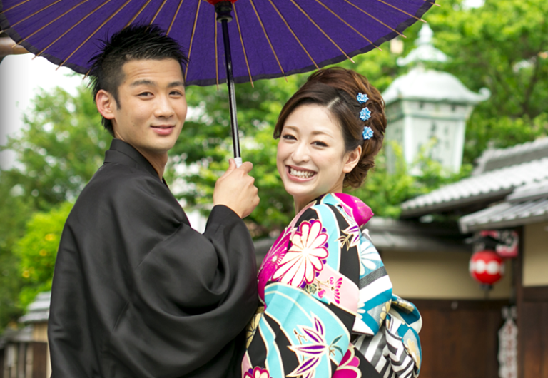 A happy Japanese couple in traditional attire pose under a purple umbrella, smiling brightly at the camera, with a lantern and greenery in the background.